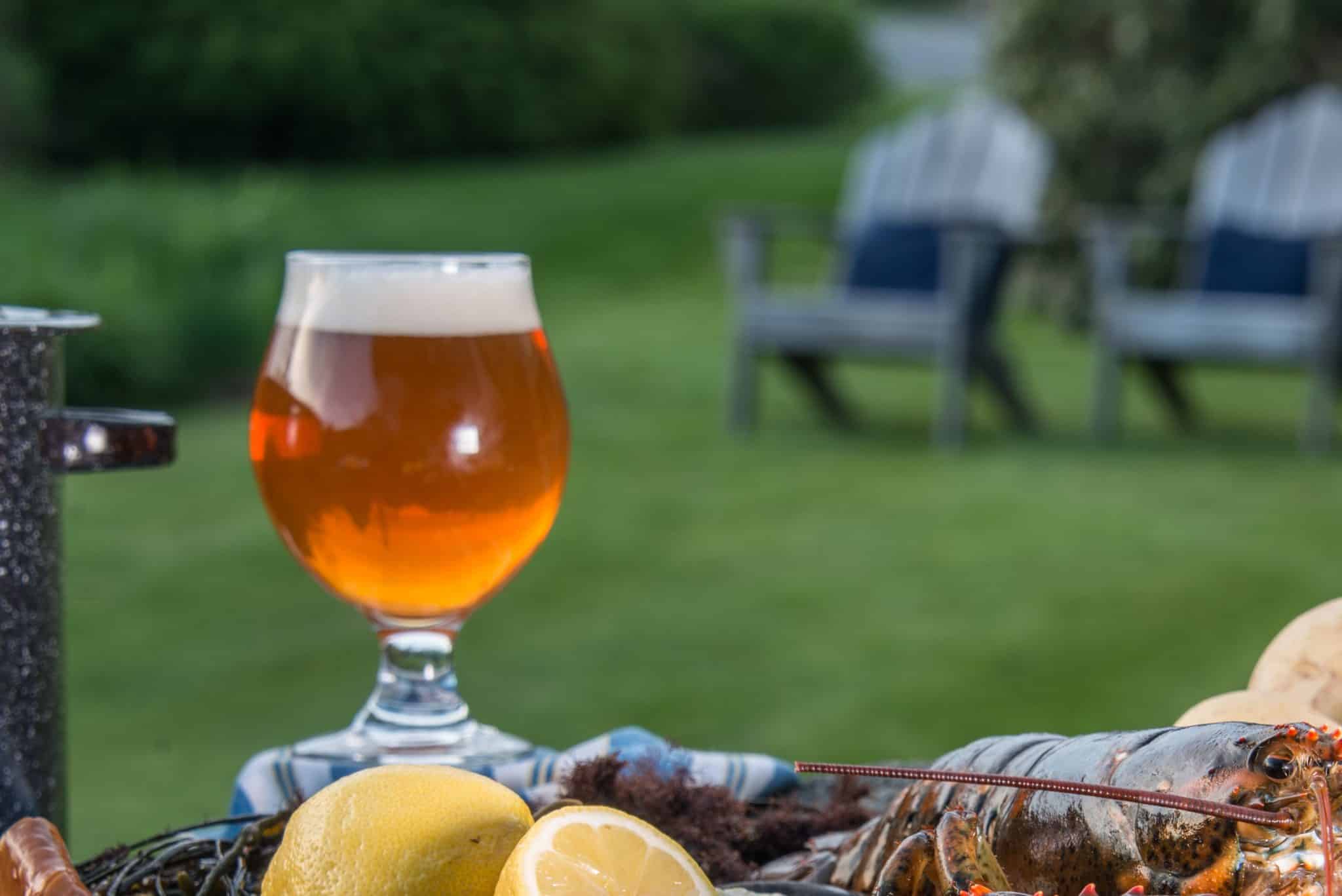 Beer in a glass outside with a lemon, lobster and chairs in the distance