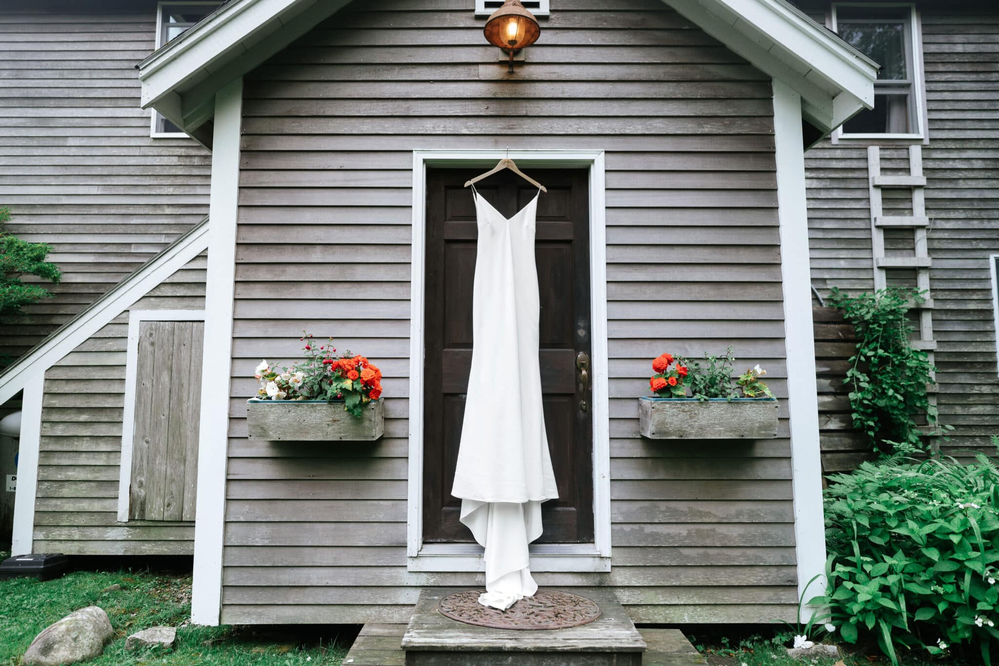 Photo of a white wedding dress hanging on an outside door, with grey weathered siding and flower boxes.
