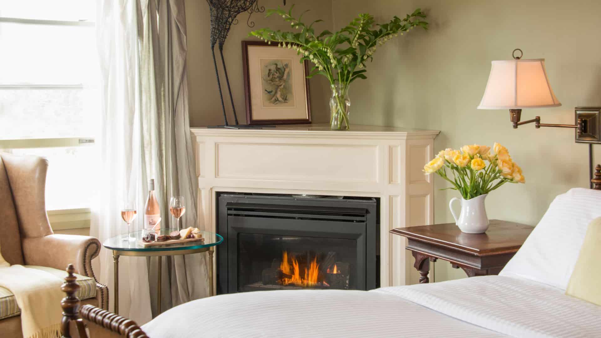 Bedroom with light green walls, white trim, corner fireplace, white bedding, wooden side table, leather chair, and small glass table with wine glasses and charcuterie board